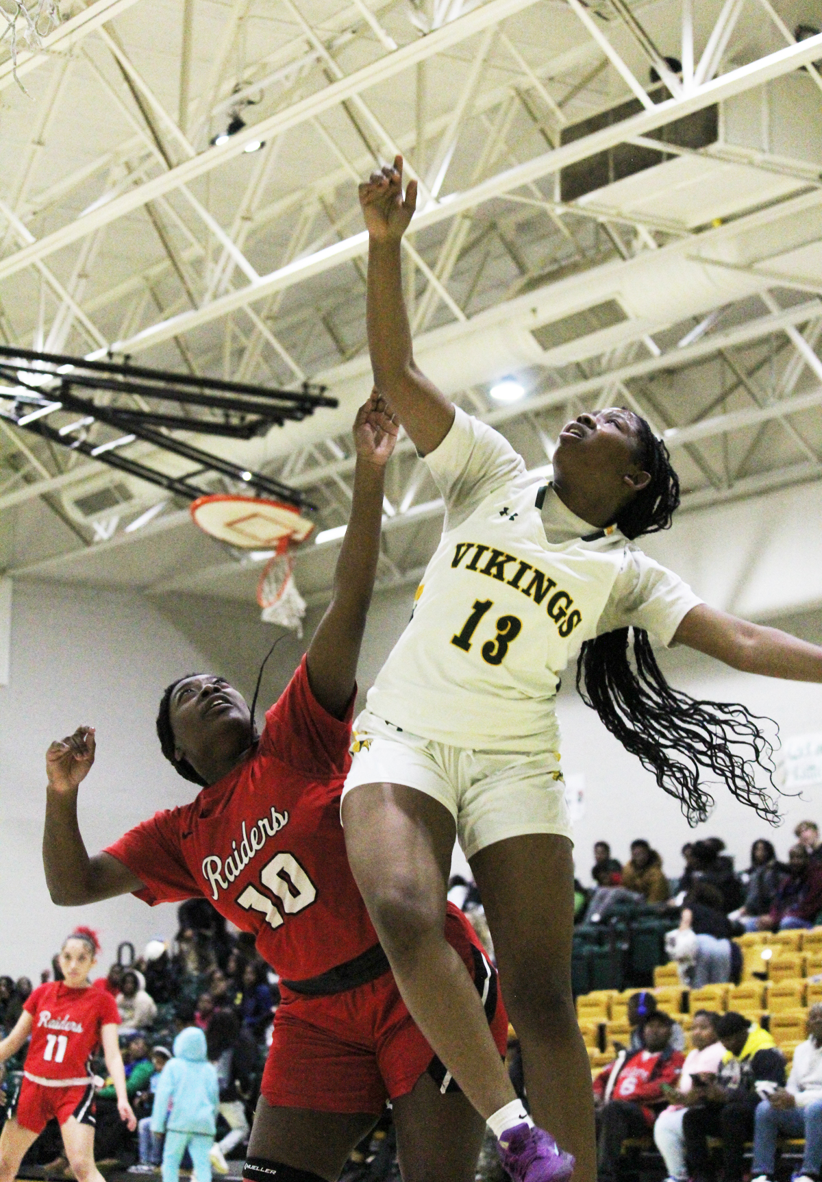Two female basketball players jump for a rebound during a game, one in a white "Vikings" uniform and the other in a red uniform, gymnasium background.
