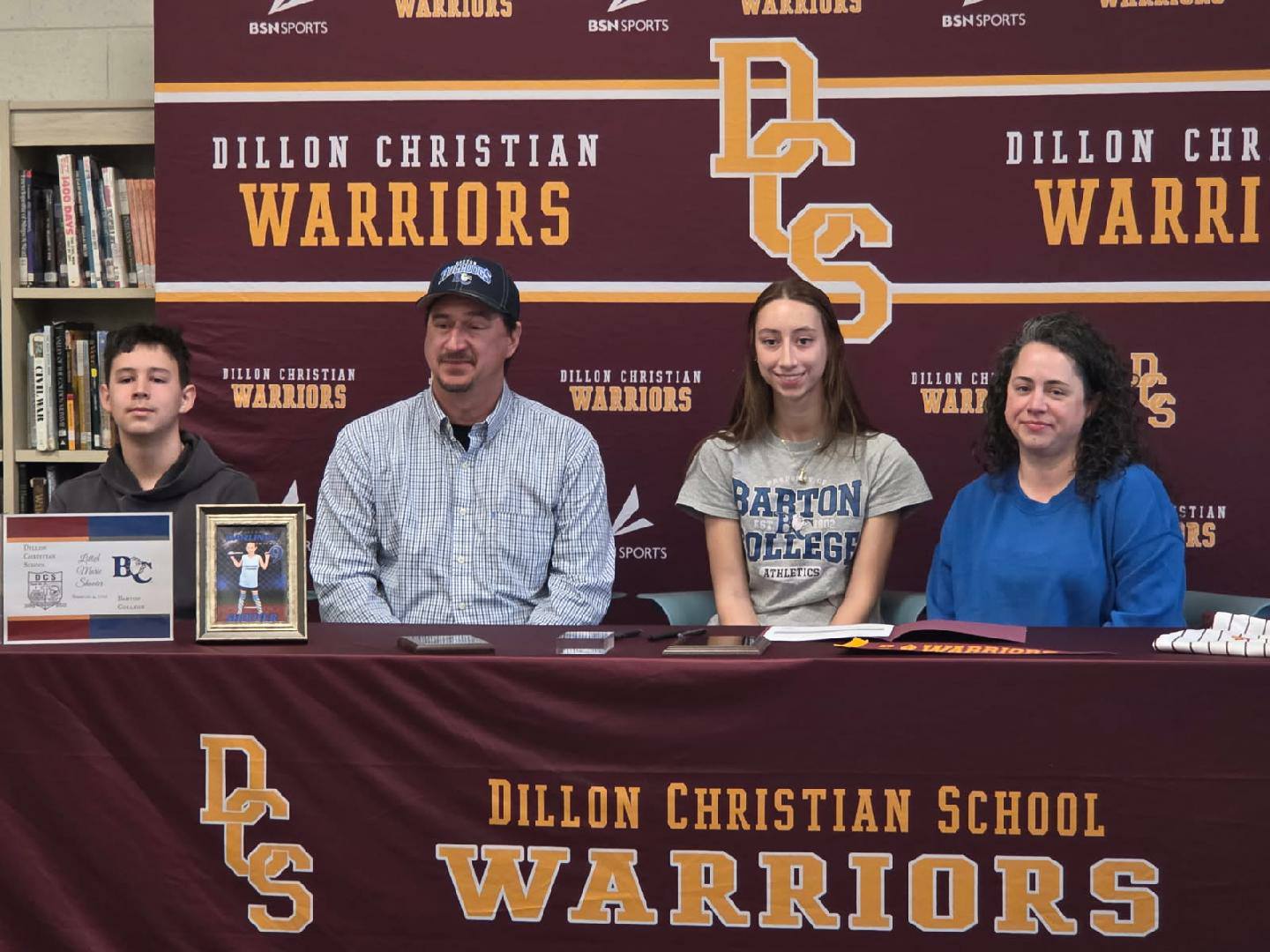 Four people sit at a table with a Dillon Christian School Warriors backdrop, posing for a photo, with one person displaying a framed picture.
