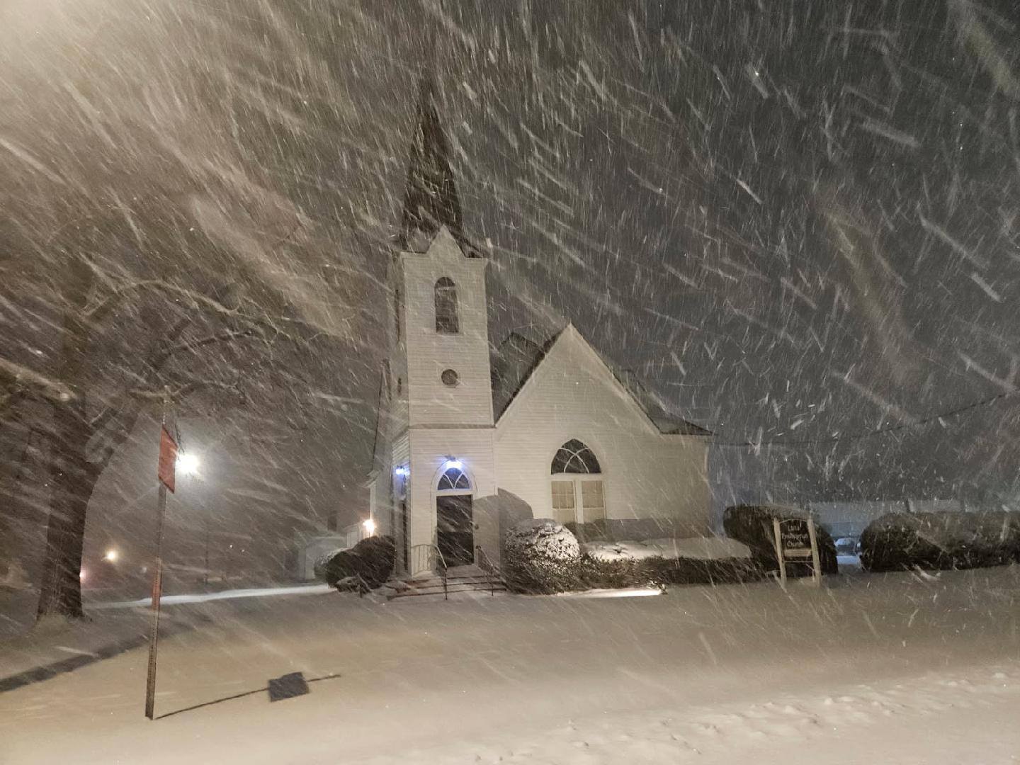 A white church with a steeple is surrounded by heavy snowfall at night, illuminated by streetlights.