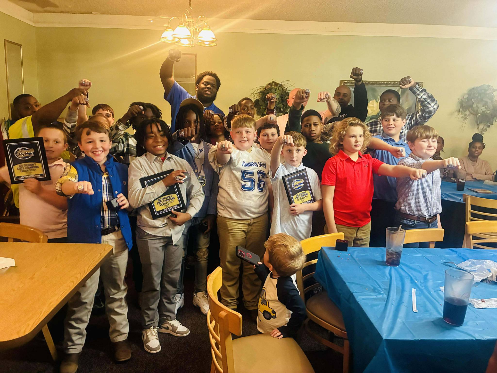 A group of children pose together in a room, some holding plaques or awards, smiling and making celebratory gestures near tables with blue tablecloths.
