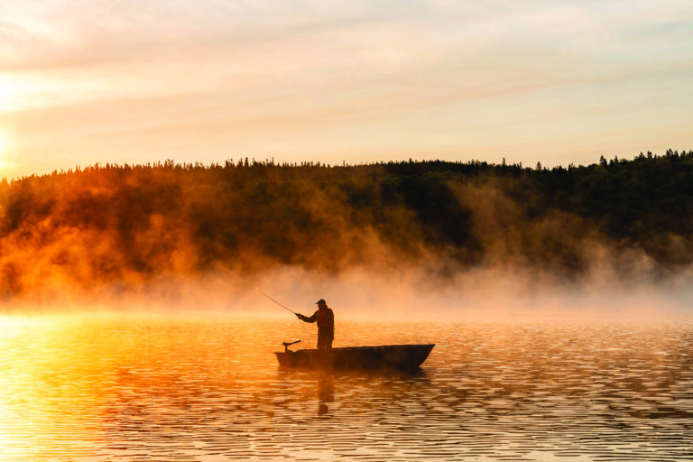 Person fishing alone in a small boat on a misty lake during sunrise, with orange light reflecting on the water and trees in the background.