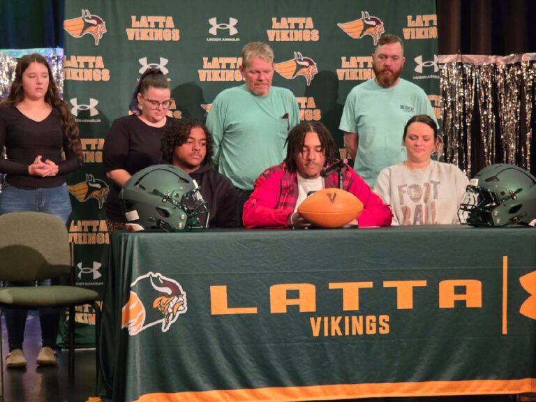 A group of people sits and stands around a table with "Latta Vikings" banner, football helmets, and a football.
