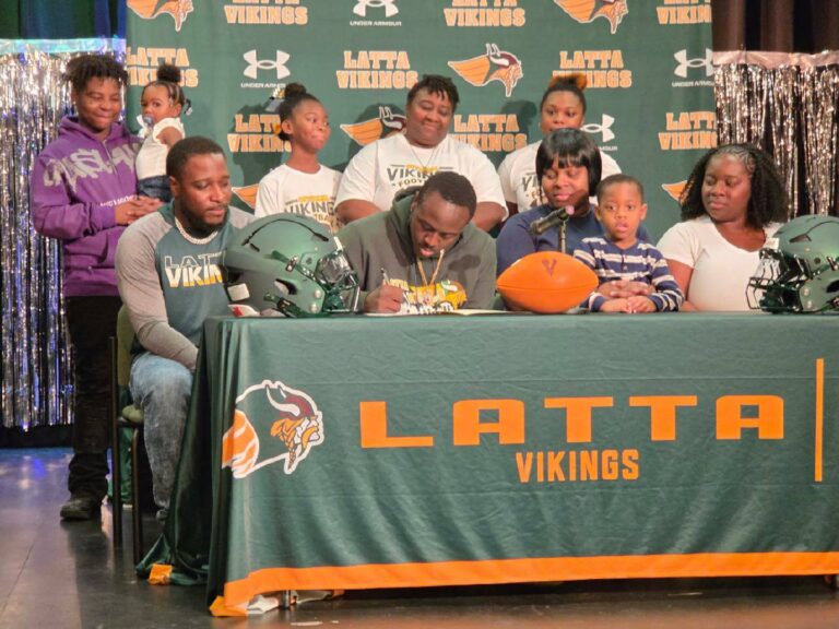 A student athlete signs a document at a table with family and teammates, under a "Latti Vikings" banner, with football gear and supporters around.