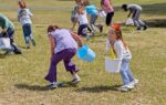 Children playing outdoors on grass, holding colorful plastic buckets, running and laughing together during a group activity on a sunny day.