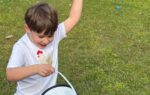 A young boy outdoors is raising his arm excitedly while holding a black-and-white bucket on a grassy lawn.