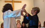 A man in a robe raises his hands toward a woman in a church, with a wooden cross and audience in the background.