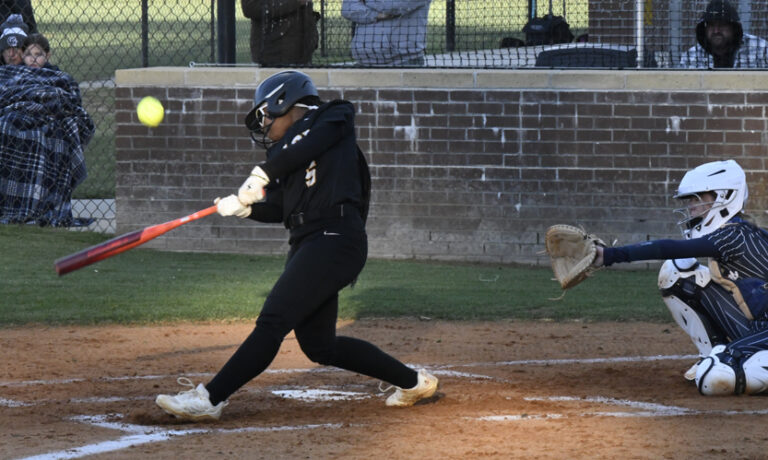 Softball player in black uniform swings bat, making contact with yellow softball during a game, as catcher prepares to catch behind home plate.