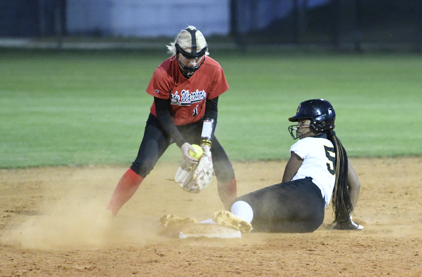A softball player in red attempts to tag out a sliding opponent in black and white on a dirt field during a game.