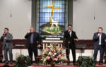 Two men singing on a church stage with stained glass window, cross, and lamb in the background, floral arrangement in front.