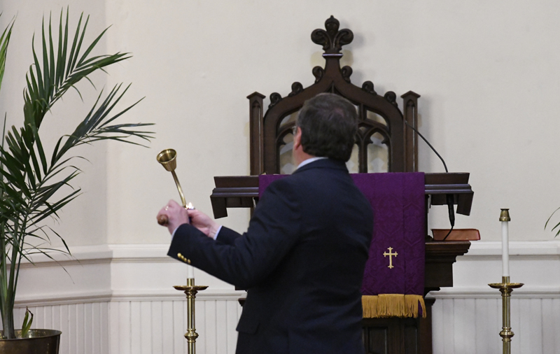 A man in a suit holding a liturgical object stands before a church altar draped in purple cloth with a gold cross, viewed from behind.
