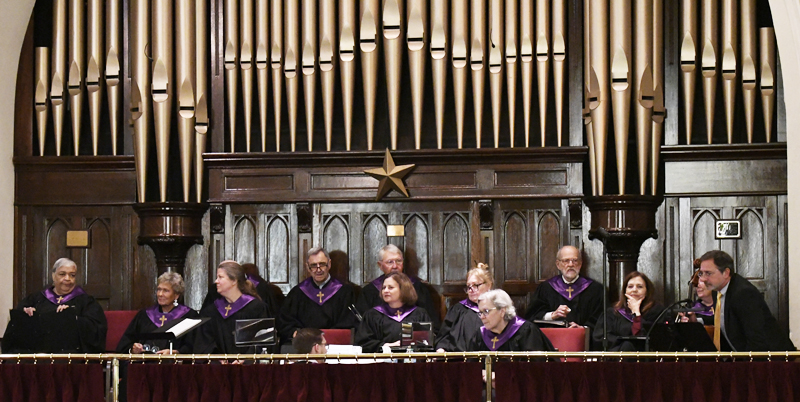 A choir of older adults in black robes with purple collars sits in front of a large pipe organ inside a church.