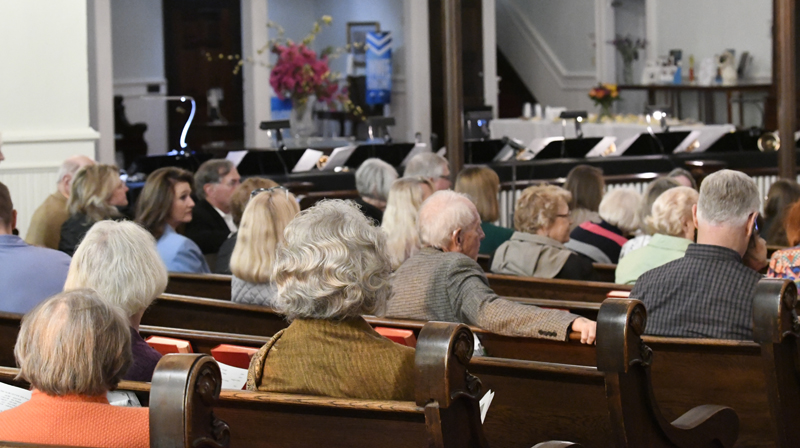 A group of people seated in church pews, facing forward toward the altar, with some floral arrangements visible in the background.