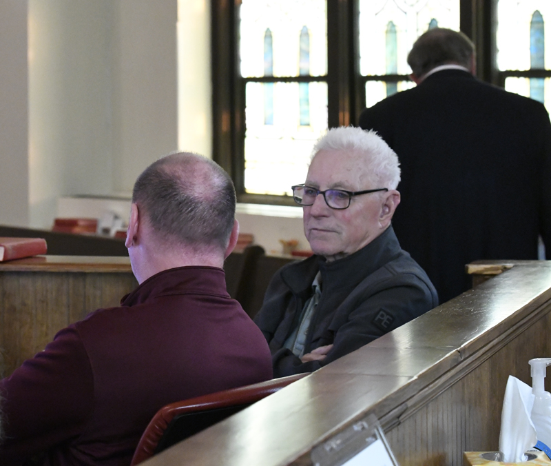 Two men sitting on a wooden pew in a well-lit room, one older man with white hair wearing glasses and a dark coat.