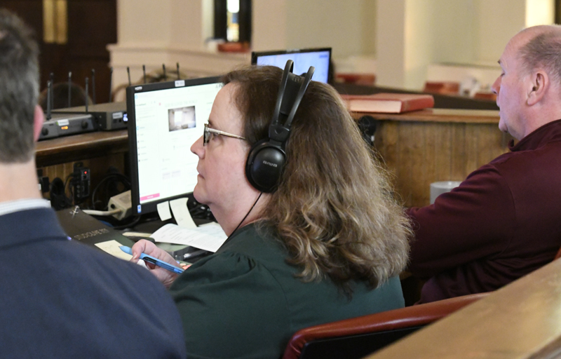 A woman with headphones sits at a desk with computers and paperwork, appearing to be focused on her work in an office or meeting environment.