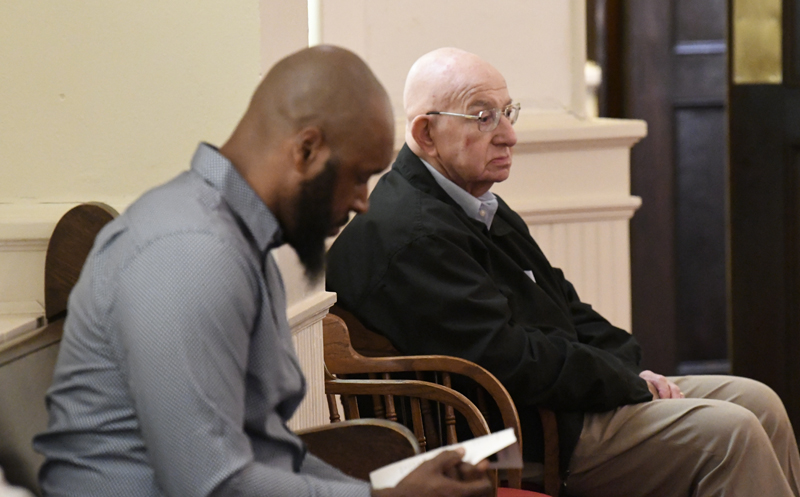Two men sit indoors, one reading a book or device, the other looking forward with a serious expression. Both are seated on wooden chairs.