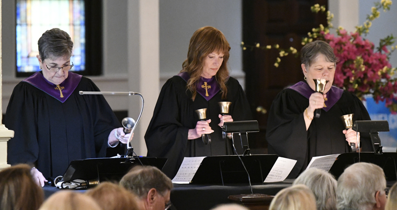 Three people in choir robes play handbells during a church performance, standing behind music stands, with an audience seated in front of them.