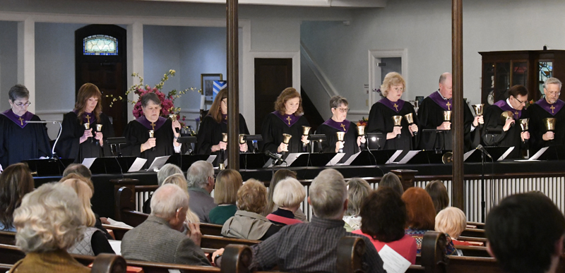 A group of women plays handbells at the front of a church while an audience watches them perform.