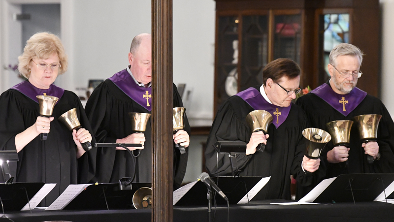Two people in black robes with purple stoles play handbells during a performance, standing behind music stands in an indoor setting.