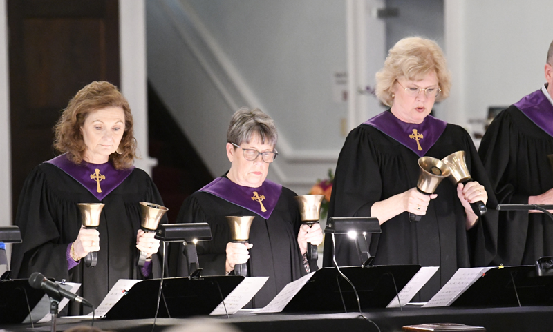 Three women in black robes with purple collars play handbells during a church performance, standing behind music stands indoors.