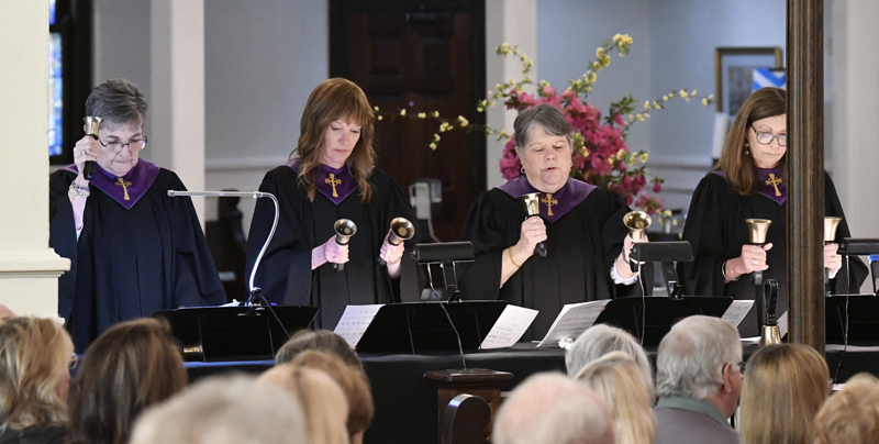 Two choir members in black robes play handbells during a church service, with sheet music in front of them and flowers in the background.