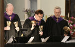 Three adults in black robes with purple stoles play handbells in a church setting, reading music sheets, with flowers visible in the background.