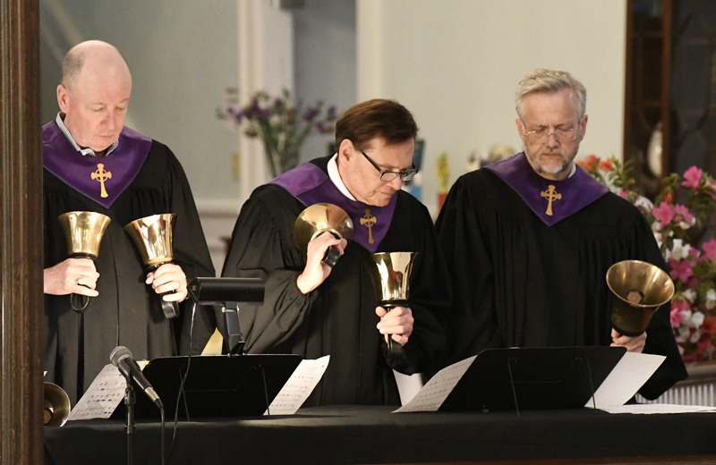 Three adults in black robes with purple stoles play handbells in a church setting, reading music sheets, with flowers visible in the background.