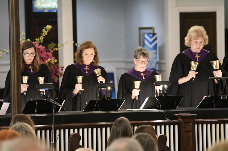 A group of women in black robes with purple collars play handbells in front of an audience inside a church.