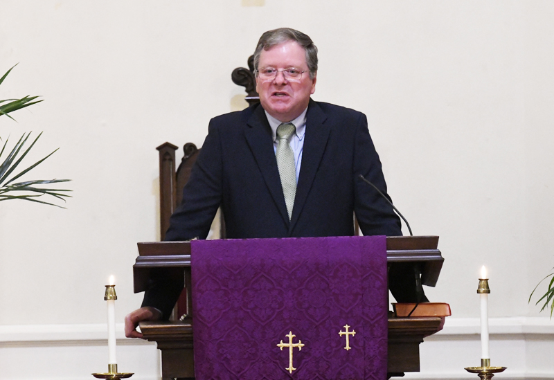 A man in a suit stands at a church pulpit adorned with a purple cloth featuring gold crosses.