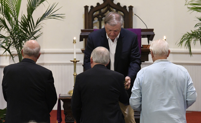 A man in a suit stands at the front of a church, greeting three older men during a ceremony or service.