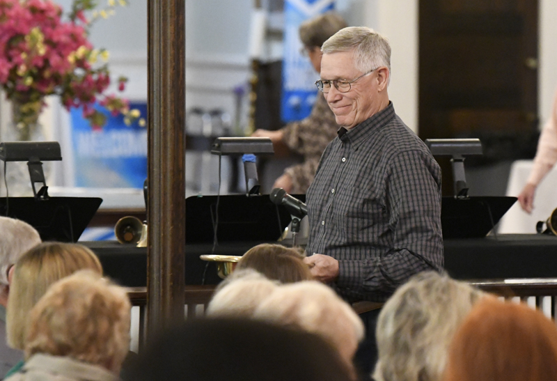 older man in glasses stands and smiles while addressing a seated audience in an indoor setting.
