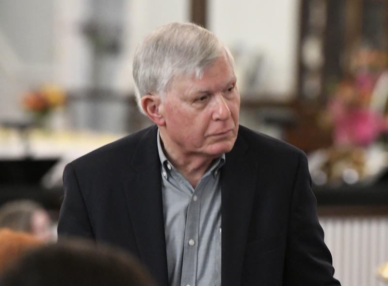 Older man with gray hair wearing a dark blazer and light-colored shirt, standing indoors and looking to the side.