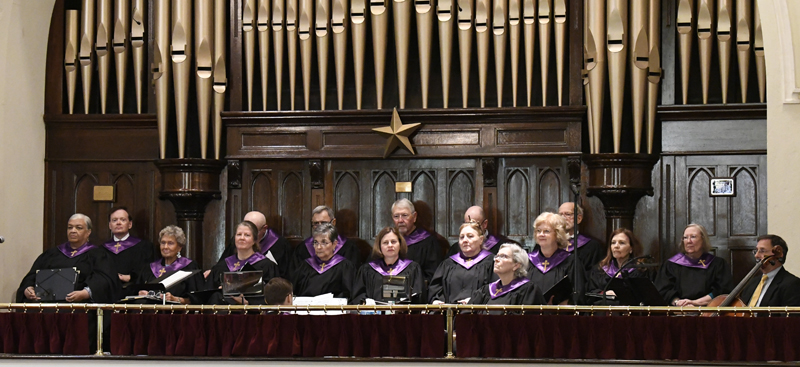 A choir of older adults in black robes with purple collars sings in front of a large pipe organ in a church.