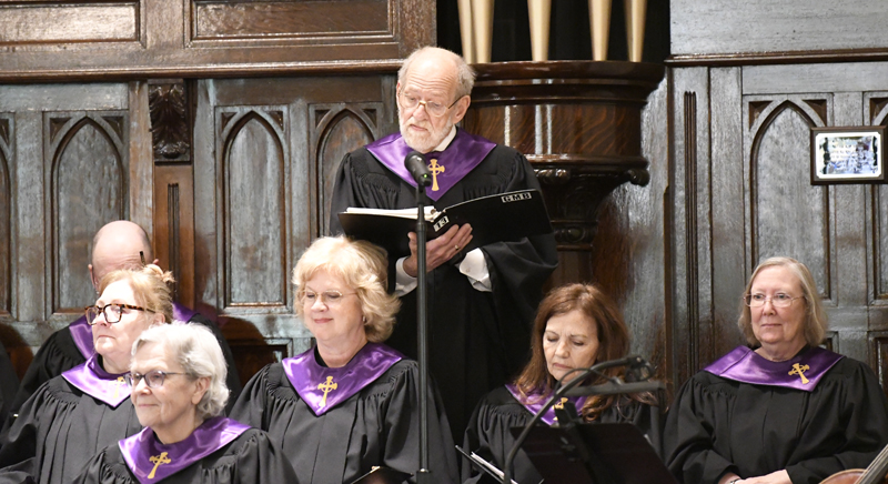 A church choir dressed in black robes with purple stoles sings, with one man standing and holding a hymnal while others are seated.