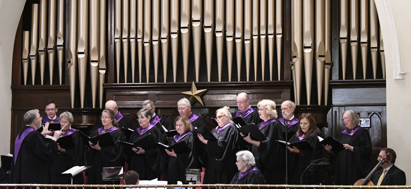 A choir of men and women in black robes with purple collars sings in front of a large pipe organ inside a church.