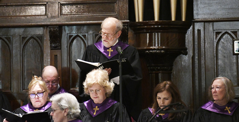 A church choir, dressed in black robes with purple stoles, sings during a performance in front of wooden panels and organ pipes.