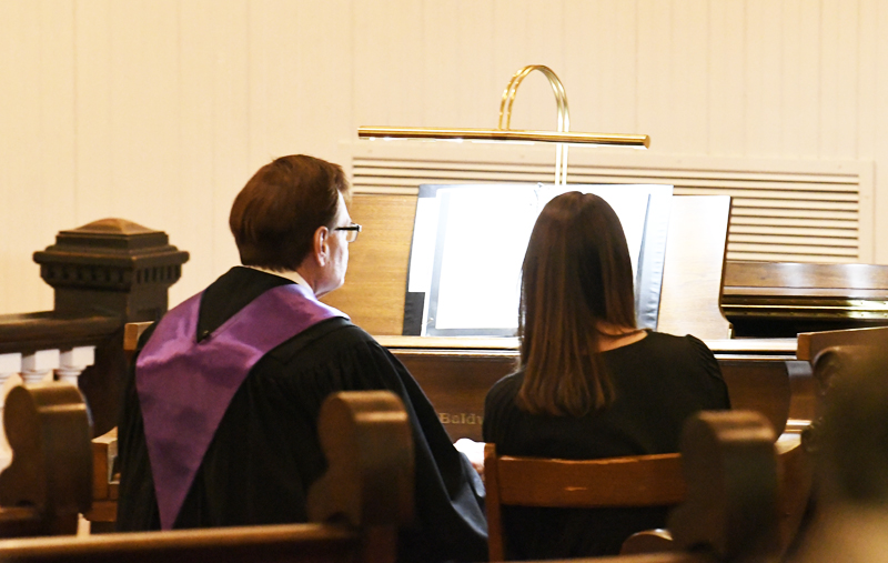 Two people sit side by side at an organ, reading sheet music, one in a black robe with a purple stole, in a softly lit room.
