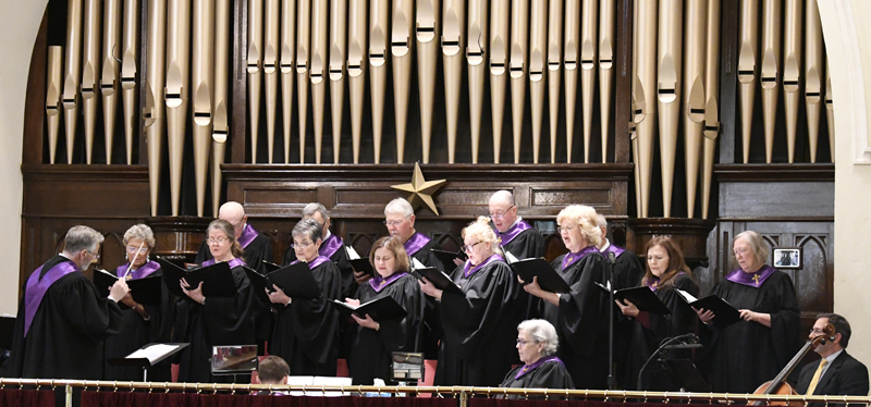 A choir of older adults in black robes with purple scarves sings in front of large organ pipes in a church setting.