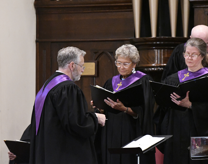 A church choir wearing black robes with purple stoles sings together while holding sheet music in a wooden interior.