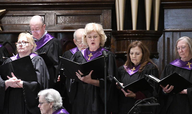 A church choir dressed in black robes with purple stoles sings together, holding sheet music, in a wooden sanctuary with organ pipes in the background.