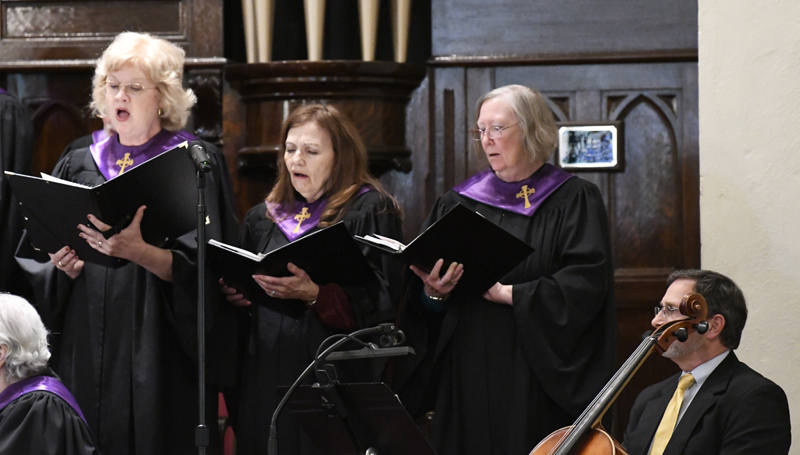 Three women in choir robes with purple collars sing from hymn books in a church setting.