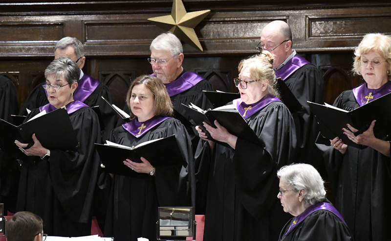 A group of adults in black choir robes with purple collars sing from hymn books in a church, standing in front of wooden panels and a gold star.