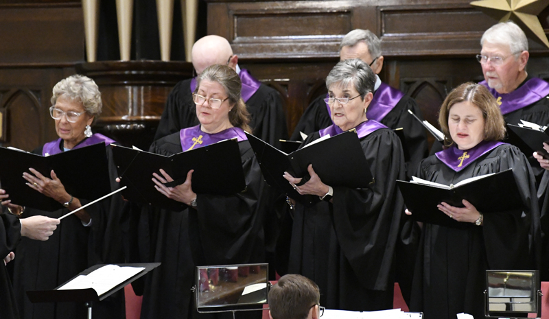 A choir of adults wearing black robes with purple stoles sings from sheet music during a performance in a church setting.