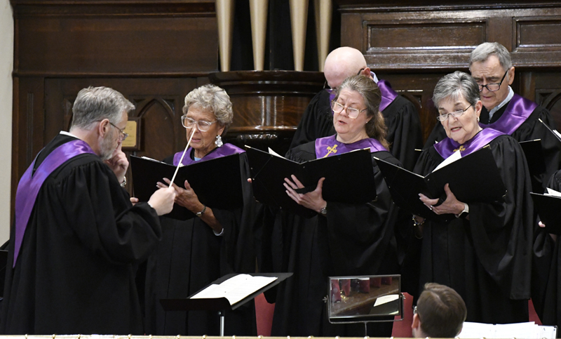 A choir dressed in black and purple sings from sheet music, led by a conductor inside a church with organ pipes in the background.
