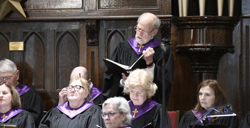 A group of choir members in black robes with purple collars sing, with one man standing and reading from a hymnal book.