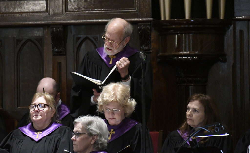 A choir of men and women in black robes with purple collars sing from hymn books inside a church.