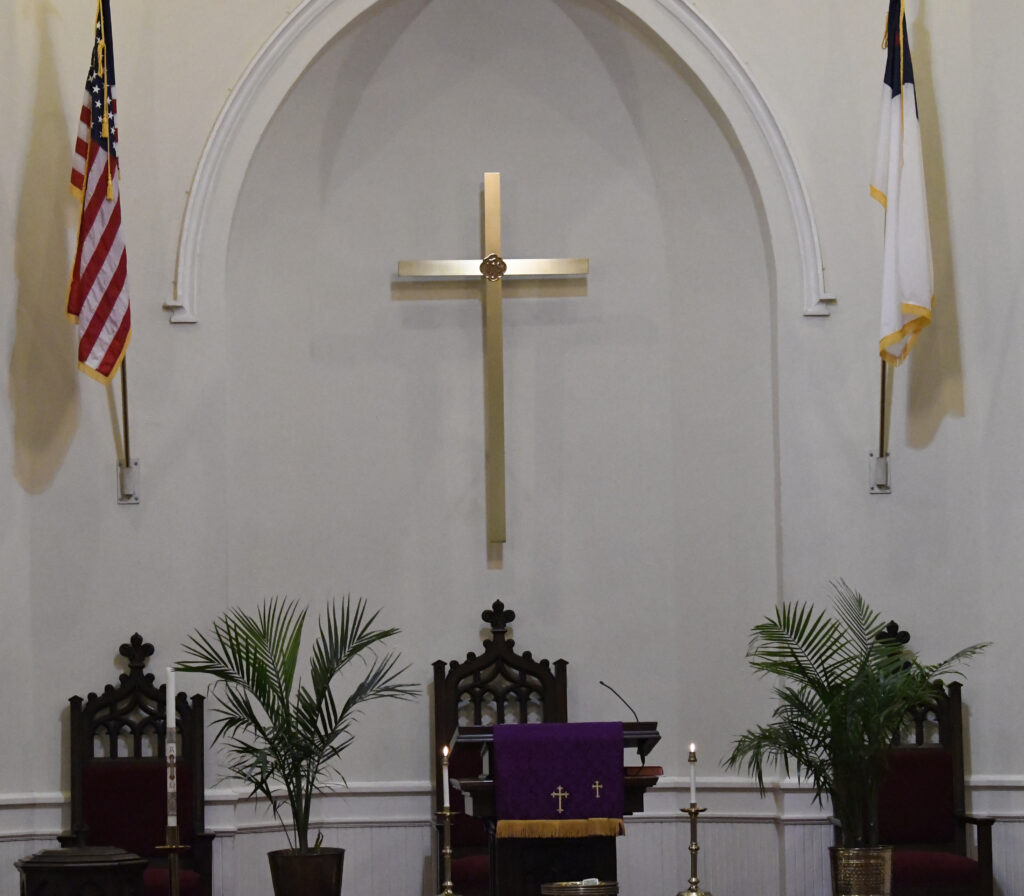 A church altar with a gold cross on the wall, two flags, three chairs, and potted plants on either side.