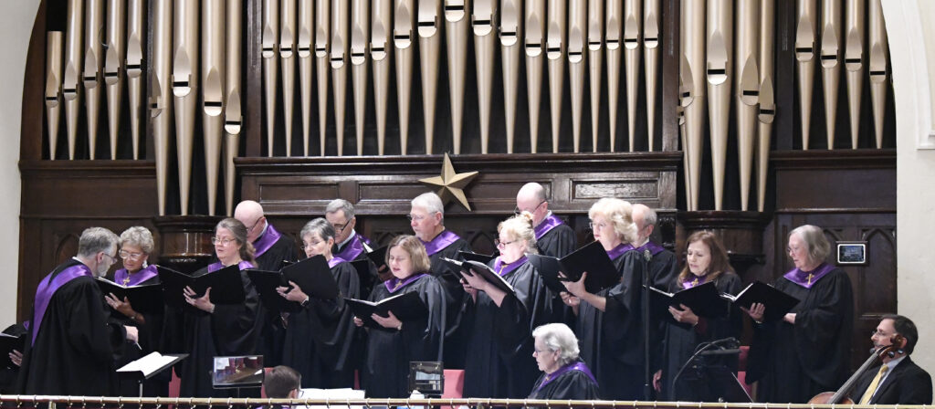 A choir group wearing black robes with purple collars sings in front of a large pipe organ in a church.