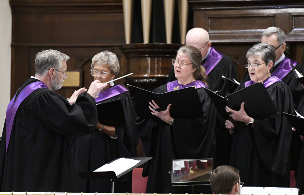 A choir dressed in black robes with purple collars sings in a church, led by a conductor.