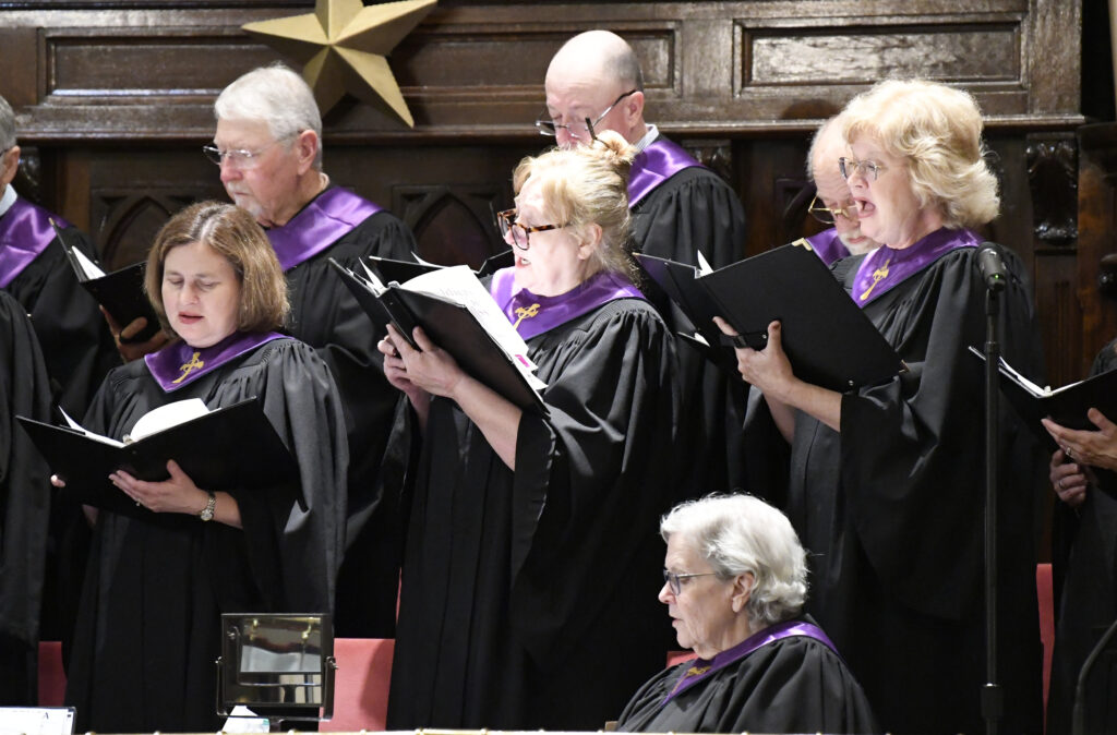 A group of choir singers in black robes with purple collars perform together, holding music books in a church setting.