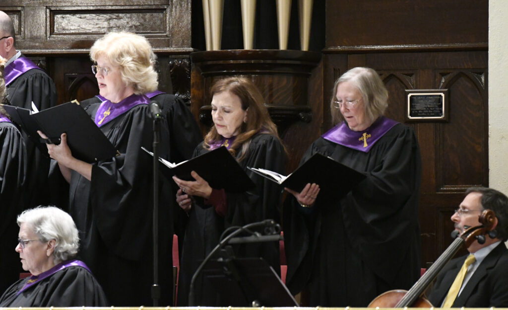 Three women in choir robes sing from hymn books during a church service, standing in front of an organ.